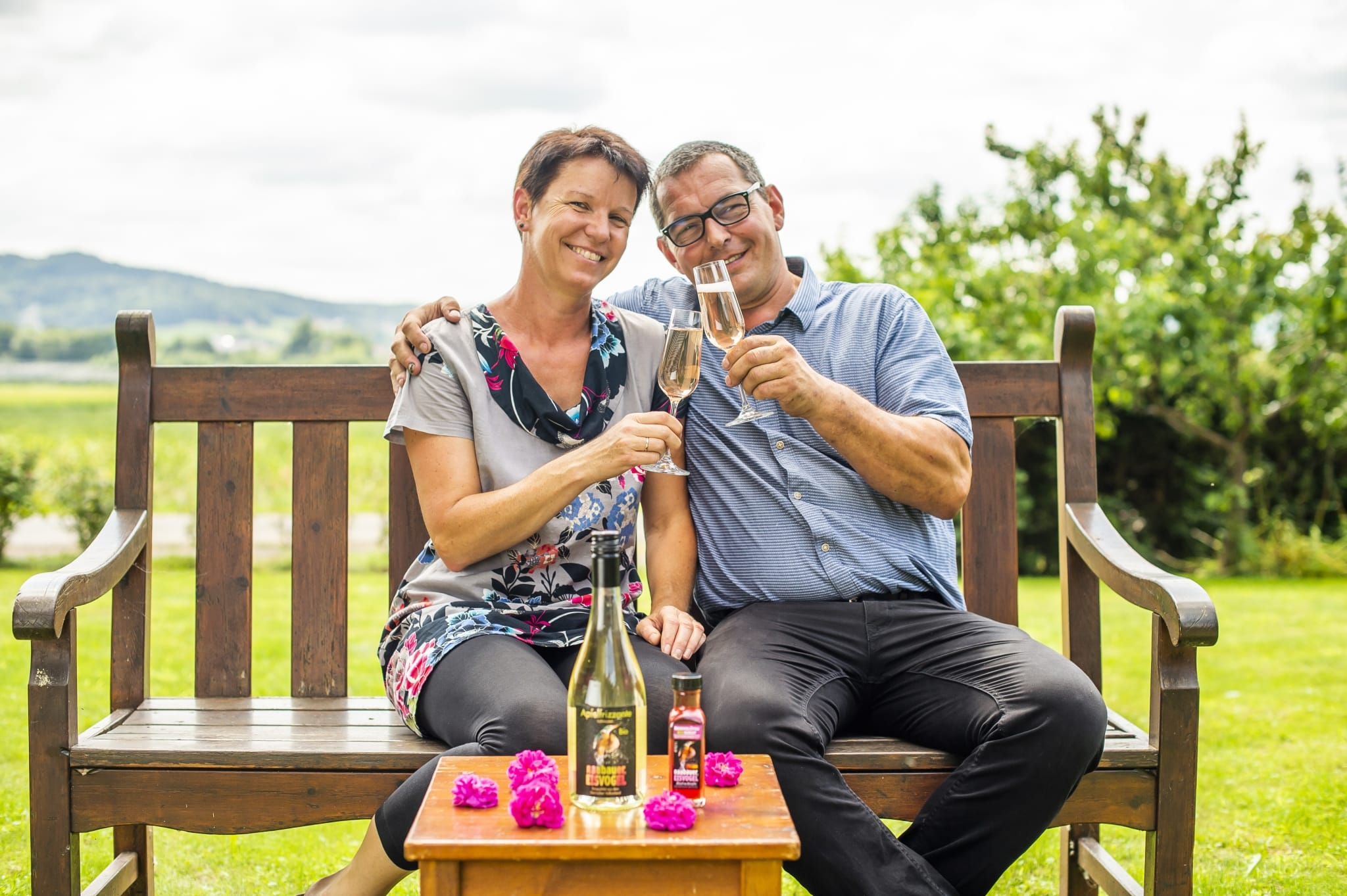 Ein Mann und eine Frau die auf einer Holzbank im Garten sitzen und ein Glas Apfelfrizzante trinken