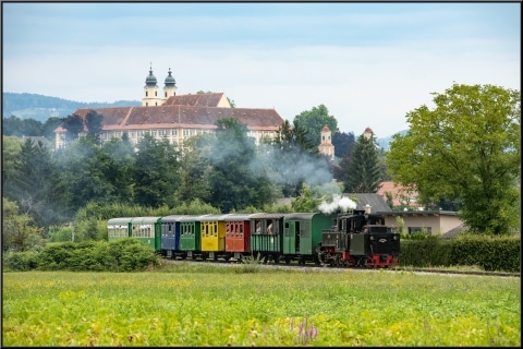 Dampfende Lokomotive fährt durch die grüne Landschaft, im Hintergrund sieht man ein Schloss