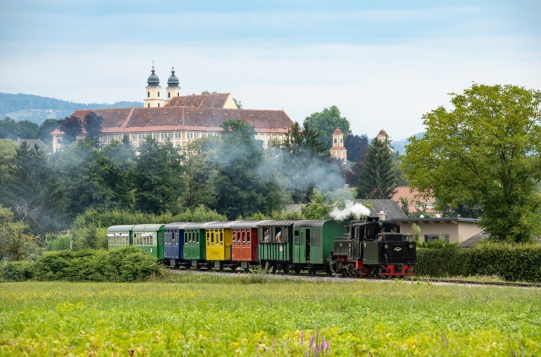 Dampfende Lokomotive fährt durch die grüne Landschaft, im Hintergrund sieht man ein Schloss