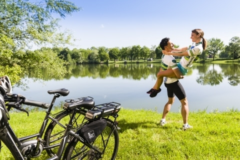 Mann in Radkleidung hält Frau in Radkleidung hoch, im Hintergrund See und grüne Wiese