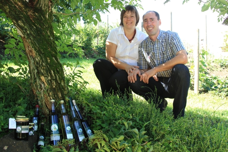 Ein Mann und eine Frau die unter einem Holunderbaum knien, im Vordergrund Holunderprodukte