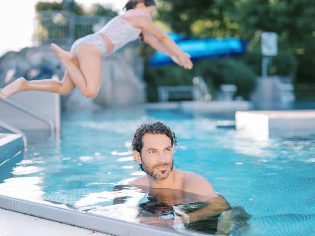 Mann schaut nach rechts und im Hintergrund springt ein Kind ins Wasser
