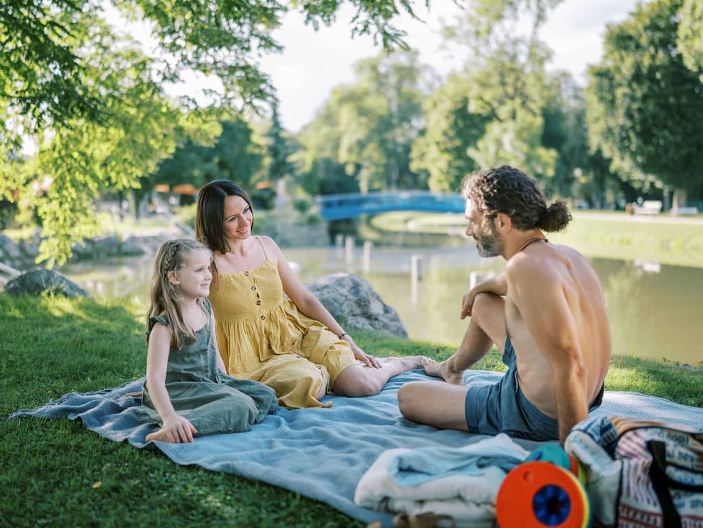 Eltern mit Tochter sitzen auf Picknickdecke vor dem Wasser