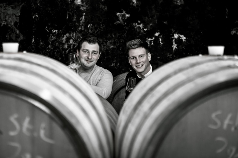 Zwei Männer mit Weingläsern, die zwischen zwei Weinfässern hervorblicken