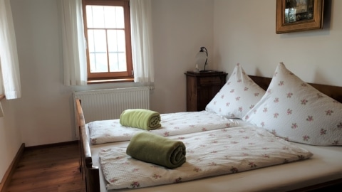 Ein Bett mit rosa geblümter weißer Bettwäsche. Auf den Decken liegen zwei grüne eingerollte Decken. Im Hintergrund ist eine dunkle Holzkommode mit einem Nachtlicht zu sehen. Das Zimmer hat weiße Wände und ein holzumrahmtes Fenster mit weißen Vorhängen.