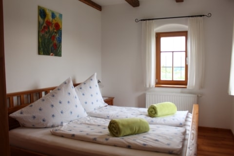 Ein Bett mit weißen Bezügen mit einem blauen Blumenmuster. Auf den Bettdecken liegen zwei eingerollte grüne Decken. Im Hintergrund ist eine weiße Wand, ein Fenster mit weißen Vorhängen und über den Kopfkissen hängt ein gemaltes Bild mit blauen und roten Blumen.