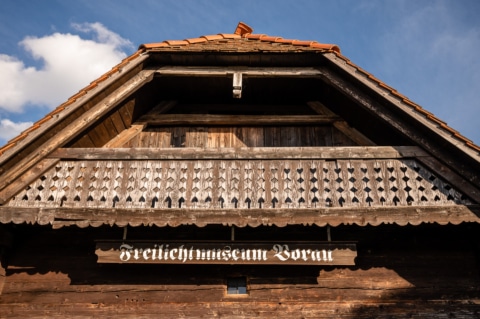 Zu sehen ist der Balkon vom Holzhaus und unter dem Balkon hängt ein Schild mit der Aufschrift "Freilichtmuseum Vorau".