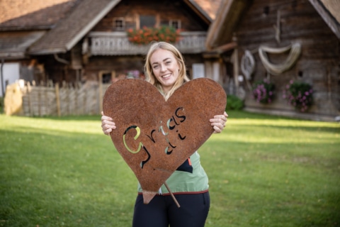 Zu sehen ist eine blonde junge Frau, sie hält ein Herz aus Metall, welches schon verrostet ist. Auf dem Herz steht "Grias di". Im Hintergrund sind die Holzhäuser zu sehen.